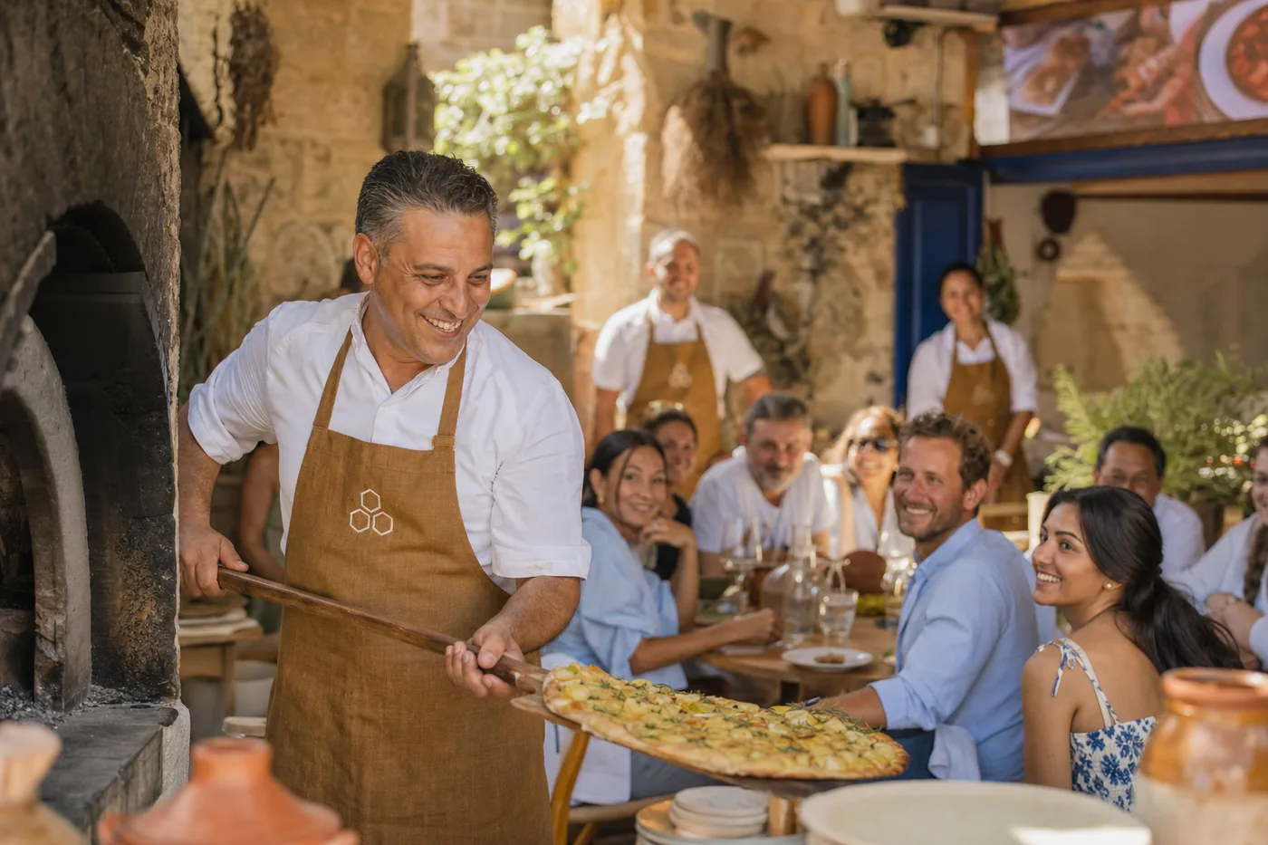 A cook slides a ftira out of the wood oven on a long peel